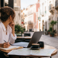 Traveler planning a trip at a cafe table with a laptop, notebook, and map
